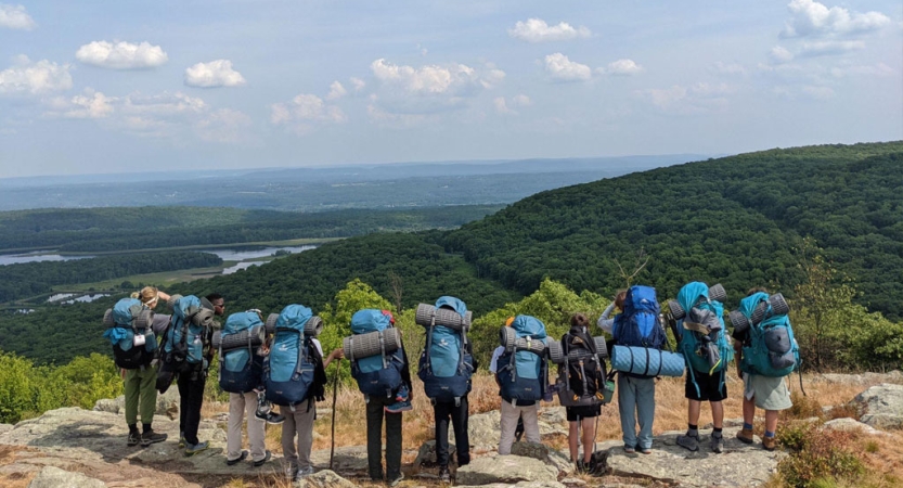 A group of people wearing backpacks stand in a line on a rocky overlook. Below them is vast green landscape, with a river winding through it. 